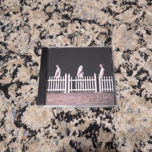Könnte beinhalten: Eine CD-Hülle mit einem dunklen Bild von drei Figuren, die hinter einem weißen Zaun stehen. Die Figuren tragen weiße Hemden. Die CD-Hülle ist schwarz.