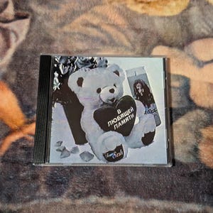 May include: A CD case featuring a black and white image. The image shows a teddy bear holding a heart with Russian text, a photo of a person, and floral elements. The text "Sesh" is also visible.