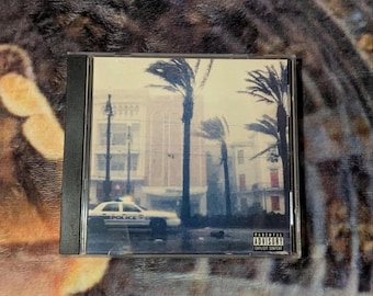 Puede incluir: Una caja de CD con un exterior de plástico negro. La portada presenta una imagen borrosa de una calle de la ciudad con palmeras y un coche de policía. El texto "Parental Advisory Explicit Content" es visible en la esquina inferior derecha.