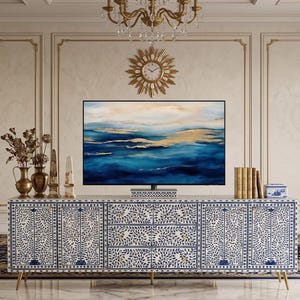 May include: A luxurious living room featuring a large blue and white patterned cabinet with gold legs, a large flat-screen TV, and two blue armchairs. The room is decorated with gold accents and a chandelier.