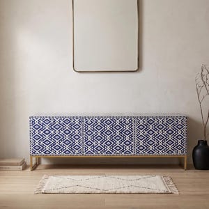 May include: A modern blue and white patterned cabinet with gold legs. A rectangular mirror hangs above the cabinet. A small rug and a black vase with branches are in front of the cabinet. The room has a neutral color palette.