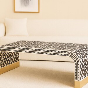 May include: A contemporary coffee table featuring a black and white geometric pattern. The table has a curved design with gold-coloured accents on the legs. The table is in front of a white sofa.