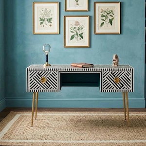 May include: A black and white geometric patterned desk with gold-coloured hardware and legs. The desk has two drawers and a central open space. Botanical prints in gold frames hang on the teal wall above the desk. A rug with a white border is on the floor.