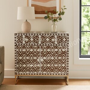 May include: A brown and white patterned cabinet with two doors and a drawer. The cabinet has a gold-colored base and a lamp on top. A sofa and a window are in the background.