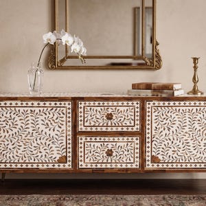Pode incluir: Aparador de madeira com um design de incrustação floral, com duas gavetas e duas portas de armário. O armário tem pernas douradas e é coberto com livros, um castiçal e um vaso com flores brancas. Um espelho com moldura dourada está pendurado acima.