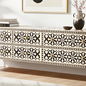 May include: A long, cream-colored dresser with six drawers, adorned with a black geometric pattern. The dresser has gold-colored knobs and legs. A vase with flowers, a small bowl, and books are on top.