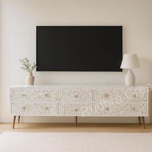 May include: A long, rectangular white sideboard featuring six drawers with intricate floral inlay patterns and small round knobs. The piece is supported by four thin, tapered legs and is positioned beneath a large, black, wall-mounted television.