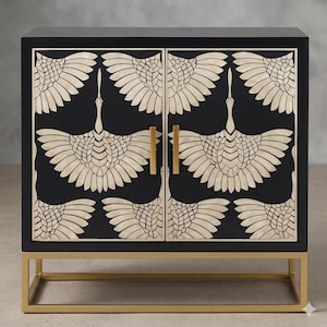 May include: Black cabinet with a gold base and hardware. The cabinet doors feature a repeating pattern of cream-colored crane designs. The cabinet is a square shape and has two doors with gold handles.
