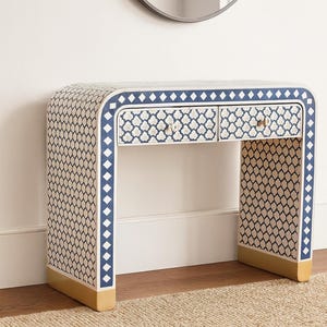 Può includere: Una scrivania decorativa dal design curvo. La scrivania presenta uno sfondo color crema con un motivo ripetuto di forme blu navy e bianche. I bordi sono rifiniti con diamanti blu navy. La scrivania ha due cassetti e dettagli dorati.