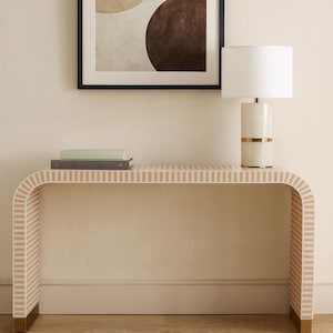 Puede incluir: Una consola con un patrón de rayas crema y beige, coronada con una lámpara blanca con una banda dorada y una obra de arte abstracta enmarcada. La mesa tiene patas doradas y está contra una pared color crema.
