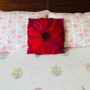 May include: A white bed with a red floral pillow and two white pillowcases with pink floral patterns. The bedspread has a white background with small green and yellow floral designs.