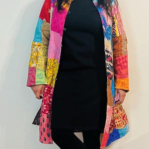 May include: A long, colorful patchwork jacket with a button closure. The jacket is made of different fabrics and patterns, including floral, geometric, and paisley designs. The jacket is worn over a black mock neck shirt and black leggings.