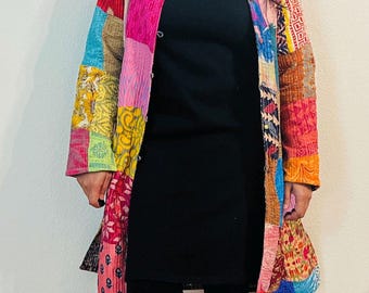 Può includere: Una lunga giacca patchwork colorata con chiusura a bottone. La giacca è realizzata con diversi tessuti e motivi, tra cui disegni floreali, geometrici e paisley. La giacca è indossata sopra una maglietta nera a collo alto e leggings neri.