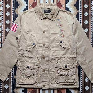 May include: Tan jacket with multiple pockets, button closures, and an embroidered design on the front. Features an American flag patch on the left sleeve. The jacket has a collared neckline and a quilted lining.