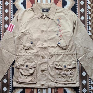 May include: A beige utility jacket with a small American flag patch on the left sleeve. The jacket has a button closure, two large flap pockets on the front, and a small embroidered design on the right side. The jacket is made of a durable cotton fabric.