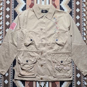 Peut inclure: Une veste utilitaire beige avec un petit patch de drapeau américain sur la manche gauche. La veste a un motif de broderie florale sur le côté droit et plusieurs poches.