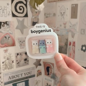 May include: A white sticker with the text "This is boygenius" and a picture of three fuzzy, pastel-colored kittens in a basket.