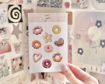 May include: A sheet of donut-shaped stickers in various colours and designs, including pink, yellow, and brown. The stickers are held in a hand against a backdrop of other decorative items. The text "bloom sweet" is printed on the top of the sticker sheet.