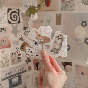 May include: A set of four stickers featuring a brown rabbit, a fawn, a gray kitten, and a pink heart. The stickers are on a white background and have a vintage aesthetic.