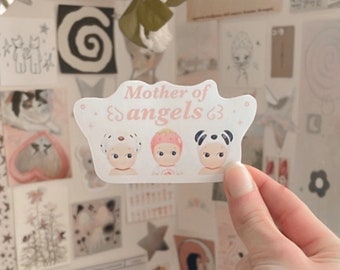 May include: A white sticker with pink text that reads "Mother of angels." The sticker features three cartoon babies wearing different hats. One baby is wearing a white shower cap, one is wearing a pink strawberry hat, and one is wearing a black and white panda hat.