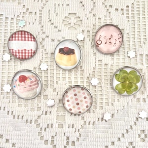 May include: Six circular magnets with silver edges, each featuring a different design. The designs include a red and white checked pattern, a custard dessert with a cherry, a cupcake, musical notes, a polka dot pattern, and a four-leaf clover.