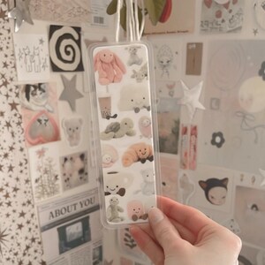 May include: A clear plastic bookmark with a variety of colorful plush toys printed on it. The bookmark features a pink bunny, a cloud, a turtle, a brown bear, a cat, and a cup with a face.