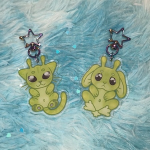 Op de afbeelding: Twee sleutelhangers met cartoonachtige wezens in lichtgroen. Eén lijkt op een kat, de ander op een konijn, beide met grote ogen en stervormige sluitingen. De achtergrond is een zachte, lichtblauwe stof.