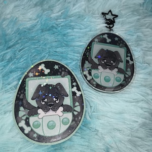 May include: Two egg-shaped acrylic charms featuring a black puppy illustration inside a light green digital pet device frame. The design includes white bone icons and holographic star accents on a dark background. One charm has a black star clasp.