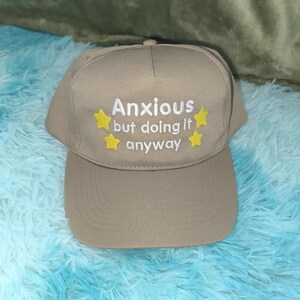 May include: A tan baseball cap featuring white embroidered text that reads Anxious but doing it anyway, accented by four small yellow embroidered stars positioned around the lettering on the front panel of the hat.
