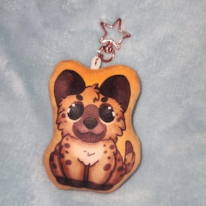 May include: A plush keychain with a cartoon hyena design. The plush has a tan body with brown spots and dark brown ears. It has large, expressive eyes and a rose gold star-shaped clip.
