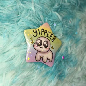 May include: A star-shaped pin with a cartoon character and the word "YIPPEE!". The pin has a gradient background of yellow, pink, and purple. The cartoon character has large eyes and a small mouth. The pin is on a blue and turquoise fluffy surface.