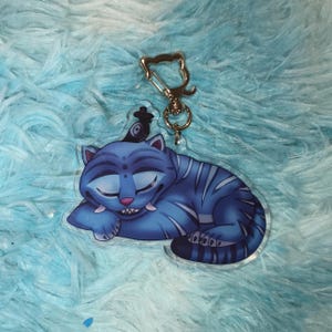 May include: A blue tiger keychain with a silver clasp. The tiger is sleeping with its eyes closed and has blue stripes. The keychain is made of clear acrylic and has a small black hat on its head.