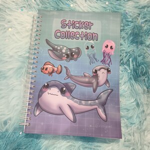 May include: A spiral-bound notebook with a cover that reads "Sticker Collection." The cover features cartoon illustrations of various sea creatures, including sharks, stingrays, jellyfish, and a clownfish. The background is a gradient of blue and purple.