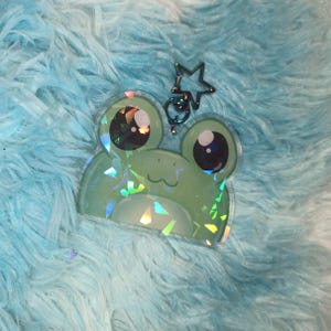 May include: A light green frog-shaped keychain with large, iridescent eyes and a smiling mouth. The keychain has a star-shaped clasp and a small ring. The background is a fluffy, light blue material.