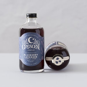 Op de afbeelding: Twee glazen flessen bosbessen-lavendel siroop. De flessen bevatten een donkerpaarse vloeistof en zijn gelabeld met "Crescent Simples". E&eacute;n fles staat rechtop, de andere ligt op zijn kant. Het etiket luidt "Bosbessen Lavendel Simple Syrup, Vers Fruit & Kruiden, netto 236ml."