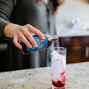 Op de afbeelding: Een helder glas is gevuld met ijs en een donkerrode vloeistof wordt uit een fles gegoten. De fles heeft een blauw etiket met het woord "Crescent" en de woorden "City" en "Berry Shrub". De drank wordt bereid op een gespikkelde aanrecht.