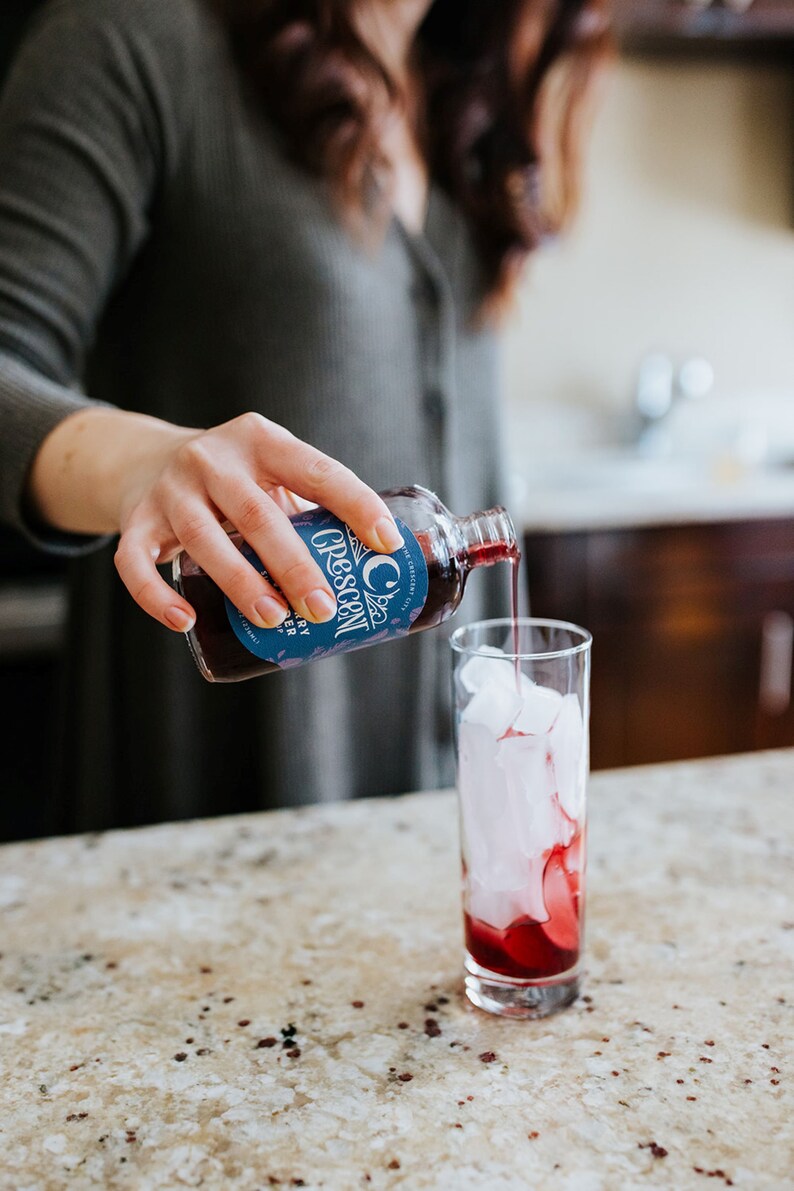 Op de afbeelding: Een helder glas is gevuld met ijs en een donkerrode vloeistof wordt uit een fles gegoten. De fles heeft een blauw etiket met het woord "Crescent" en de woorden "City" en "Berry Shrub". De drank wordt bereid op een gespikkelde aanrecht.