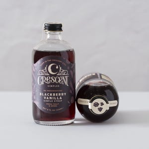 May include: Two glass bottles of Blackberry Vanilla Simple Syrup. The bottles have a dark purple label with the text "Crescent Simples" and "Blackberry Vanilla Simple Syrup". The bottles contain a dark red liquid and are 8 fl oz (236ml).