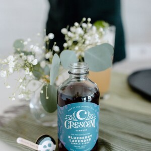 Op de afbeelding: Een heldere glazen fles Crescent Simples Blueberry Lavender Simple Syrup, met een blauw en wit etiket. De fles staat op een groen gestreepte doek, met een kleine maatlepel. Op het etiket staat "Born in the Crescent City".