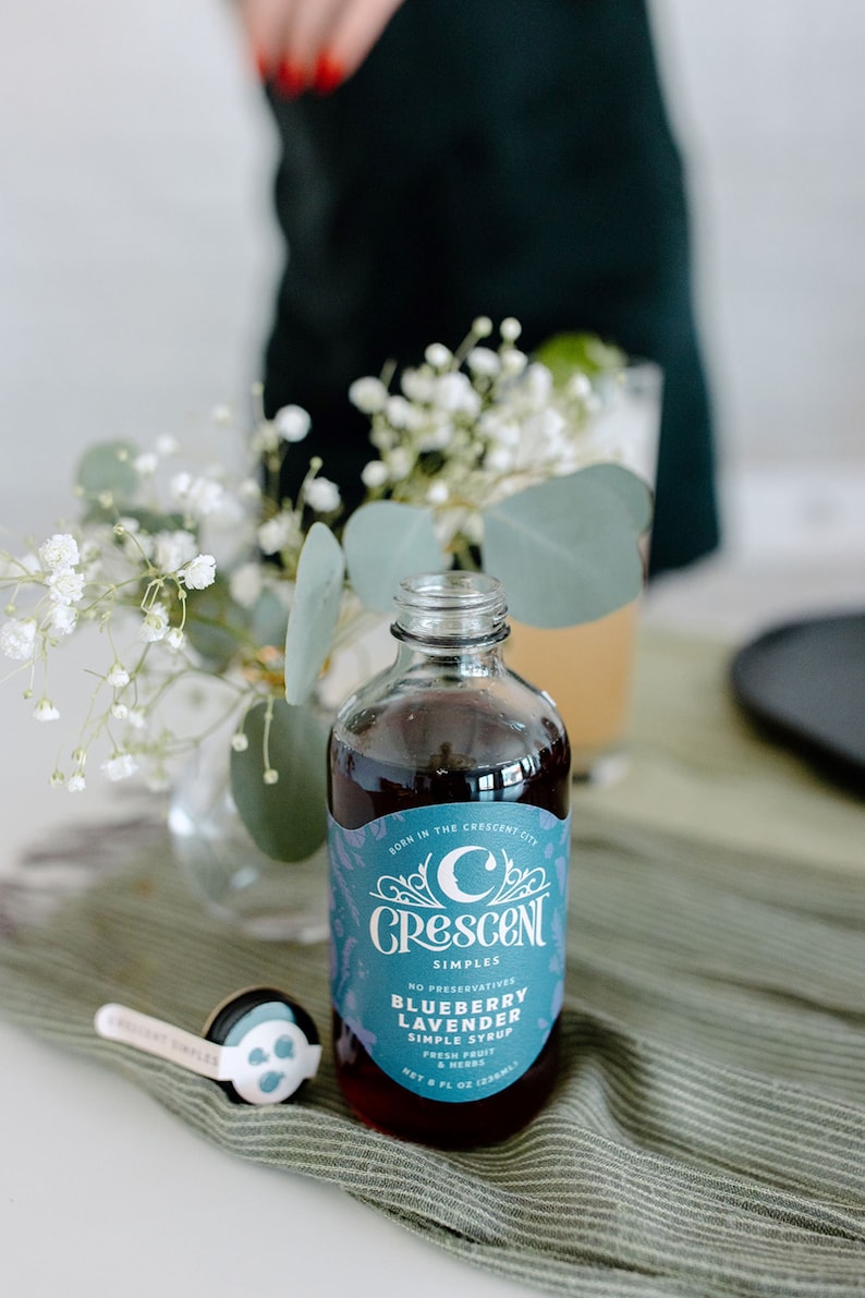 Op de afbeelding: Een heldere glazen fles Crescent Simples Blueberry Lavender Simple Syrup, met een blauw en wit etiket. De fles staat op een groen gestreepte doek, met een kleine maatlepel. Op het etiket staat "Born in the Crescent City".