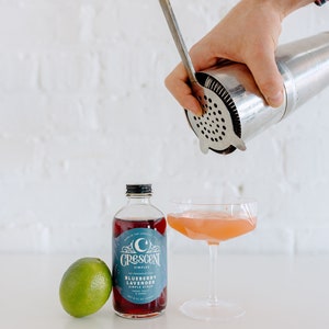 Op de afbeelding: Een cocktail wordt in een coupeglas gegoten. Een fles Crescent Simples Blueberry Lavender siroop en een limoen staan naast het glas. Een cocktailshaker en een barlepel worden gebruikt.