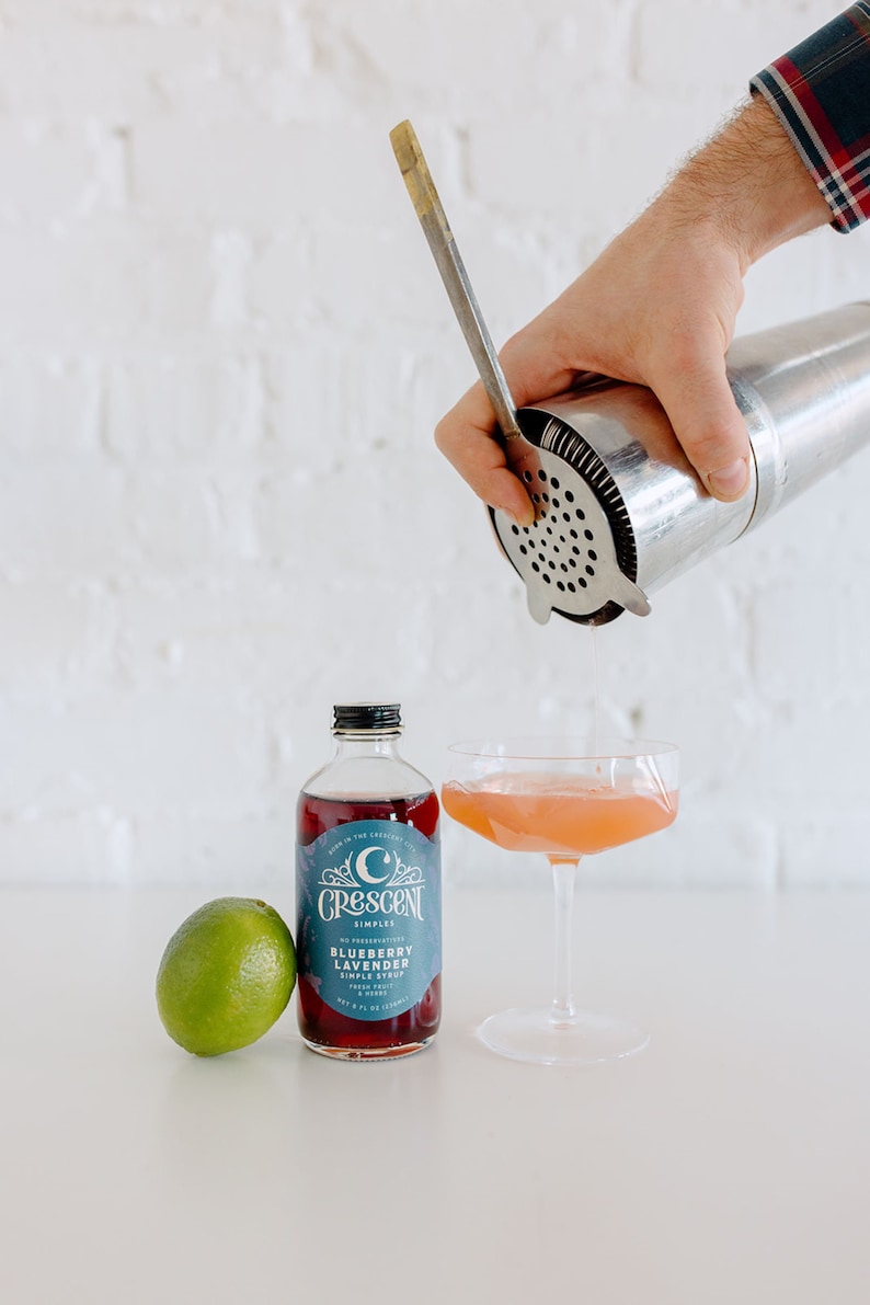 Op de afbeelding: Een cocktail wordt in een coupeglas gegoten. Een fles Crescent Simples Blueberry Lavender siroop en een limoen staan naast het glas. Een cocktailshaker en een barlepel worden gebruikt.