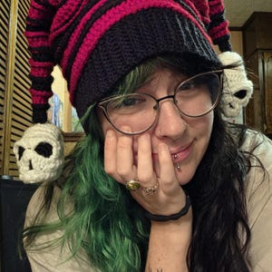 May include: A crocheted jester-style hat featuring horizontal stripes in black and dark pink. The hat has two long, pointed ends, each tipped with a small, white crocheted skull featuring black yarn detailing for the eyes and nose.