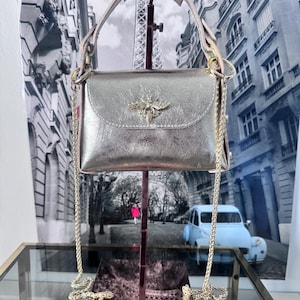 Puede incluir: Un bolso de mano plateado metalizado con un cuerpo rectangular estructurado y cierre de solapa. Un emblema de abeja en tono dorado está centrado en la solapa. El bolso incluye un asa superior corta y una correa larga y fina de cadena dorada.
