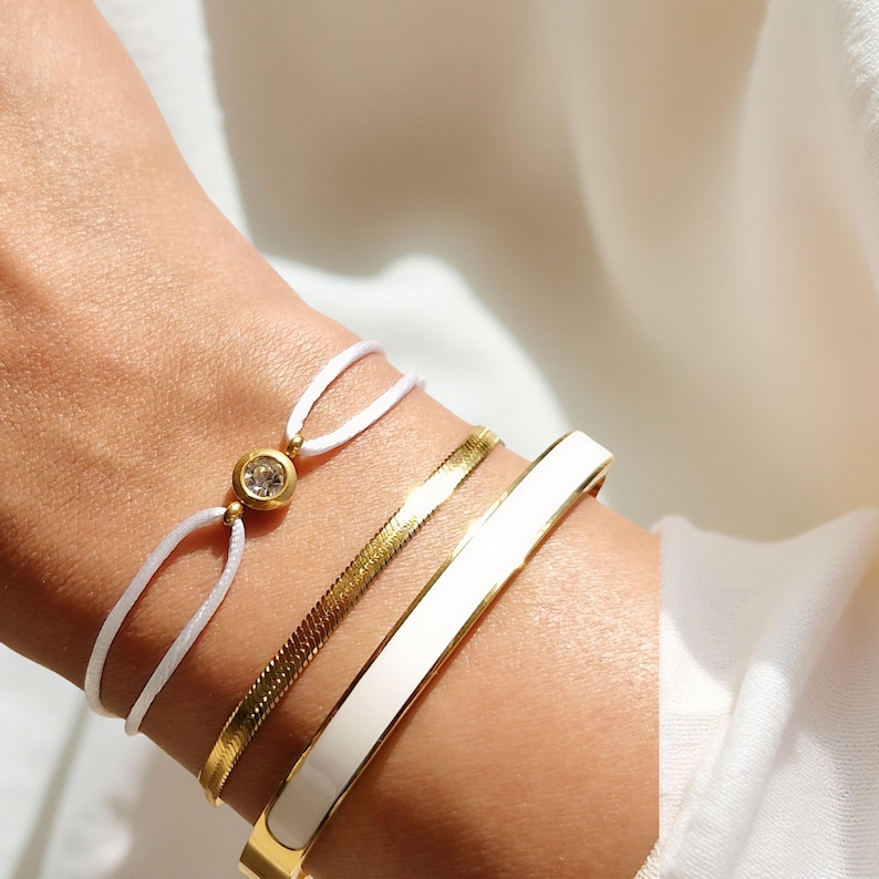 May include: Three gold bracelets on a wrist. The top bracelet is a white string bracelet with a small gold charm with a clear stone. The middle bracelet is a gold chain bracelet with a textured pattern. The bottom bracelet is a gold bangle with a white enamel finish.