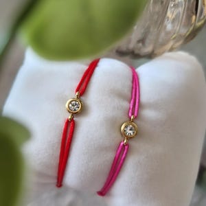 May include: Two red and pink string bracelets with gold accents and clear crystal stones.