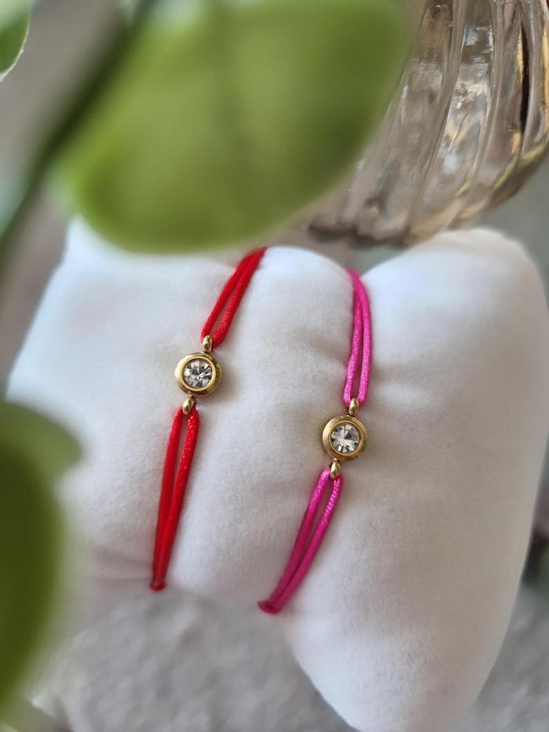 May include: Two red and pink string bracelets with gold accents and clear crystal stones.