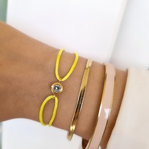 May include: A yellow string bracelet with a clear rhinestone accent. The bracelet is worn with a gold chain bracelet and a pink enamel bangle.