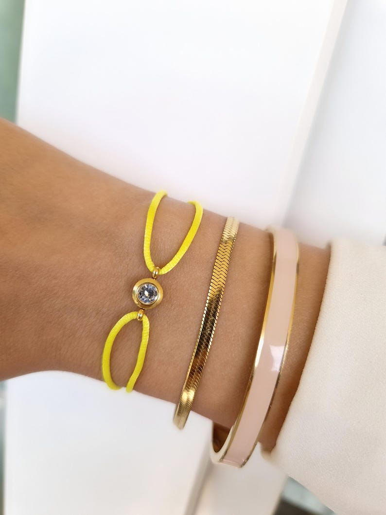 May include: A yellow string bracelet with a clear rhinestone accent. The bracelet is worn with a gold chain bracelet and a pink enamel bangle.