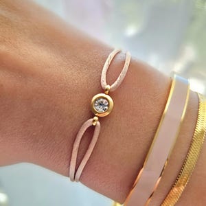 May include: A delicate pink string bracelet with a gold-toned clasp and a clear rhinestone in the center.